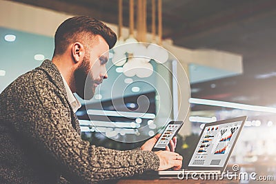 Bearded businessman sitting at table in cafe,holding smartphone and using laptop with charts,graphs on screen. Stock Photo
