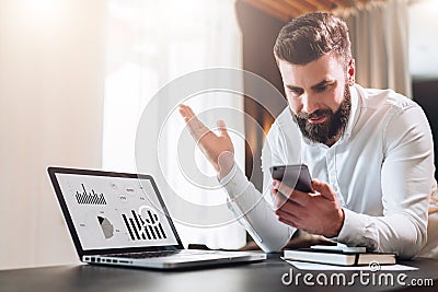 Bearded businessman in white shirt is sitting at table in front of laptop with graphs, charts, diagrams on screen Stock Photo