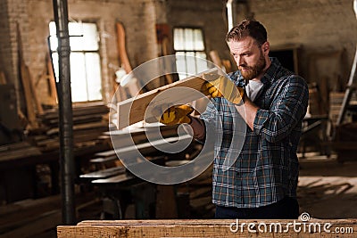 bearded carpenter in protective gloves checking wooden plank Stock Photo