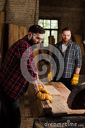 bearded carpenter in safety googles using machine saw and partner standing behind Stock Photo