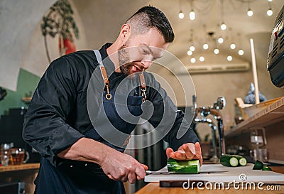 Bearded chef cook dressed black uniform with an apron chopiing fresh green cucumber with sharp knife in basement restaurant Stock Photo