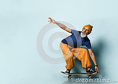 Bearded elderly man in t-shirt, sunglasses, orange pants, hat, gumshoes. Riding black skateboard, posing on blue background Stock Photo