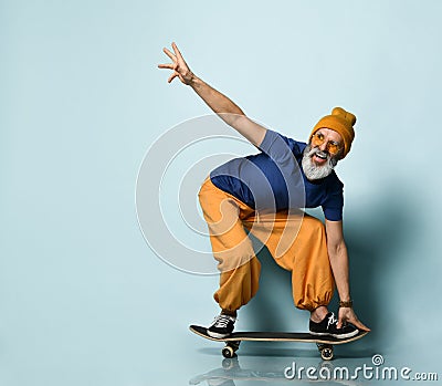 Bearded elderly man in t-shirt, sunglasses, orange pants, hat, gumshoes. Riding black skateboard, posing on blue background Stock Photo