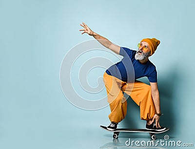 Bearded elderly man in t-shirt, sunglasses, orange pants, hat, gumshoes. Riding black skateboard, posing on blue background Stock Photo