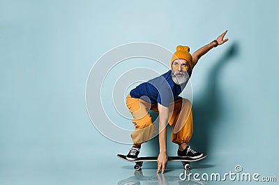 Bearded elderly man in t-shirt, sunglasses, orange pants, hat, gumshoes. Riding black skateboard, posing on blue background Stock Photo