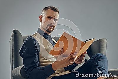 Man with stylish mustache, dressed in black shirt and trousers, beige vest is sitting on dark sofa, reading a magazine Stock Photo