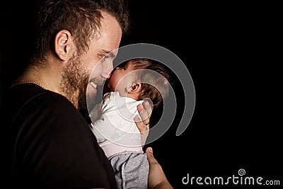 A bearded father in a black T-shirt holds a newborn baby. the child bites his fatherâ€™s nose Stock Photo
