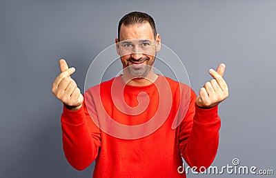 Bearded hispanic man in his 40s wearing a red sweater making the money gesture rubbing his fingers with a greedy face Stock Photo