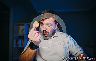 Bearded hungry man sits at night in a room with a blue light on his desk and looks at the chips in his hand with a serious face. Stock Photo