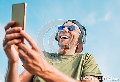 Bearded man in baseball cap, wireless headphones and blue sunglasses found something funny in smartphone device and cheerful Stock Photo