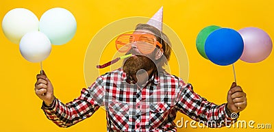 Bearded man in birthday hat and funny glasses with balloons blowing noisemaker. Party time. Holiday. Stock Photo