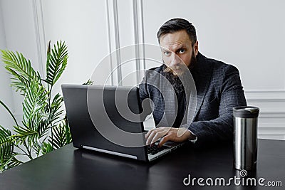 A bearded man in black golf works at a laptop at home in a bright room. Self isolation Stock Photo