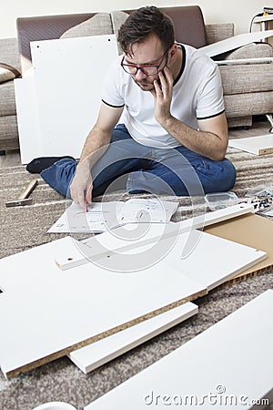 Bearded man in glasses, a white T-shirt and jeans sits on the rug thoughtfully and reads the furniture assembly instructions. Th Stock Photo