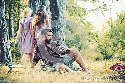 Bearded man drinking coffee or tea by campfire. Two girls in retro dresses walking on path in woods, turn back Stock Photo