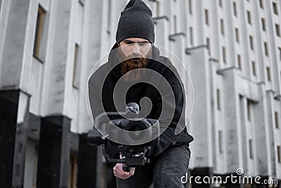 Bearded Professional videographer in black hoodie holding professional camera on 3-axis gimbal stabilizer. Filmmaker Stock Photo