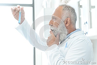 Bearded scientist in white coat thinking and looking at tube with reagent in chemical lab Stock Photo
