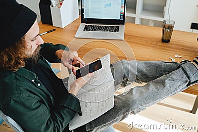 Bearded student sits with his feet up on his desk while studying online using laptop and smartphone Stock Photo