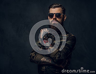 Bearded tattooed guy in military shirt and sunglasses posing with thoughtful look. Studio photo against dark wall Stock Photo