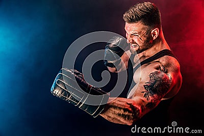 Bearded tattooed sportsman muay thai boxer in black undershirt and boxing gloves fighting on dark background with smoke. Stock Photo