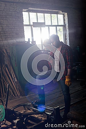 bearded worker in protective gloves wiping forehead and standing near machine Stock Photo