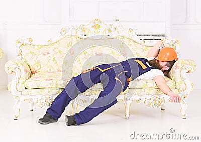 Bearded worker sleeping on vintage sofa with protective helmet over his face. Tired builder enjoying break and having Stock Photo