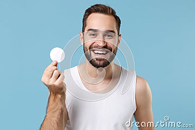 Bearded young man 20s years old in white shirt hold in hand cotton pad isolated on blue pastel wall background, studio Stock Photo
