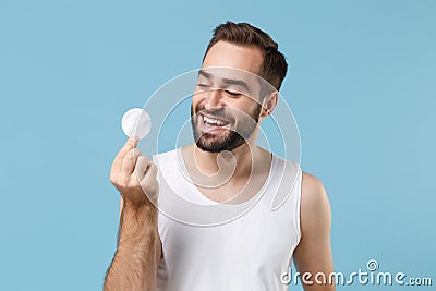 Bearded young man 20s years old in white shirt hold in hand cotton pad isolated on blue pastel wall background, studio Stock Photo