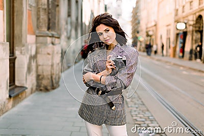 Beatiful brunette young woman in nice jacket and white pants, holding retro camera in hands. Fashion photo on urban Stock Photo