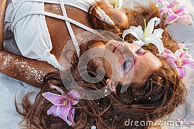 Beatiful young woman lying on sand with pink and white lilies on the beach at sunset Stock Photo