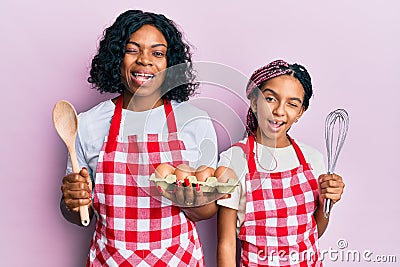 Beautiful african american mother and daughter cooking cake using baker whisk winking looking at the camera with sexy expression, Stock Photo