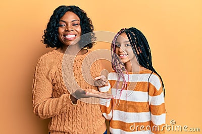 Beautiful african american mother and daughter wearing wool winter sweater amazed and smiling to the camera while presenting with Stock Photo