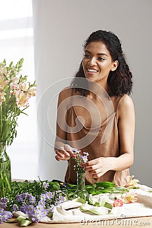 Beautiful african girl florist smiling making bouquet in flower shop over white wall. Stock Photo