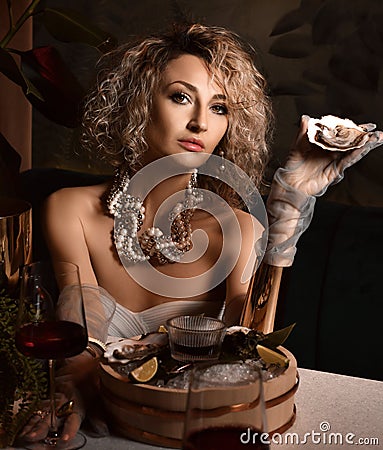 Beautiful arrogant blonde curly hair woman in seafood restaurant eating oysters and drink wine holding one in hand Stock Photo