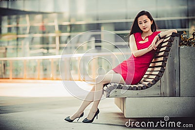 Beautiful Asian girl model in red dress sitting on a bench posing at the modern city background. Stock Photo