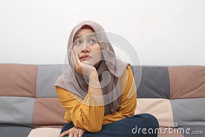 Beautiful Asian muslim woman thinking something seriously while sitting on sofa, girl with contemplation expression Stock Photo