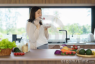 Beautiful Asian pregnant woman eating with lust cereals on breakfast in kitchen at home, enjoying meal. Yummy food for pregnant Stock Photo