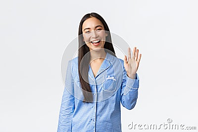 Beautiful asian woman in blue pajamas smiling happy and waving hand to say hello, greeting girlfriend and welcome to Stock Photo