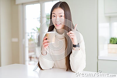 Beautiful Asian woman drinking a coffee in a take away paper cup surprised with an idea or question pointing finger with happy Stock Photo