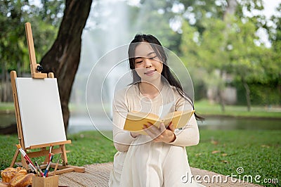 A beautiful Asian woman enjoys reading a book while picnicking in a green park on the weekend alone Stock Photo