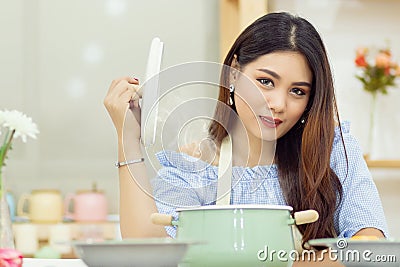 Beautiful Asian woman sit at the table in the kitchen, open the lid of the soup pot with the heat vapor Stock Photo