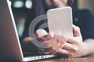 A beautiful Asian woman with smiley face holding and looking at smart phone while using laptop on wooden table Stock Photo