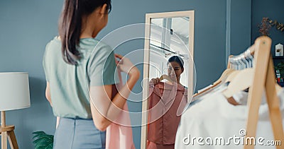 Beautiful attractive Asia lady choosing clothes on clothes rack dressing looking herself in mirror in living room at house. Girl Stock Photo