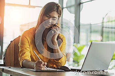 Beautiful Attractive Asian Business woman writing down requirement to book and talking with customer in smartphone Stock Photo