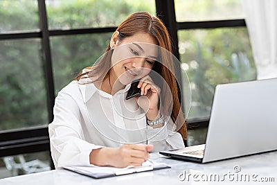 Beautiful Attractive Asian Business woman writing down requirement to book and talking with customer in smartphone Stock Photo