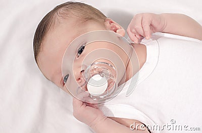 Beautiful baby lying down on white sheets with a pacifier in his mouth. Stock Photo