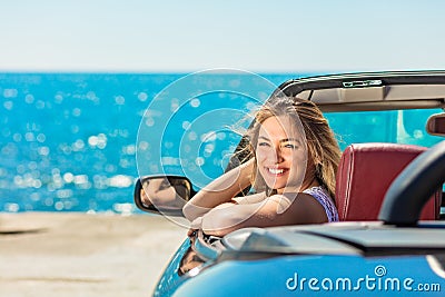 Beautiful blond smiling young woman in convertible top automobile looking sideways while parked near ocean waterfront Stock Photo