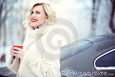 Beautiful blond woman wearing luxurious white fur coat drinking hot coffee on snowy winter day and laughing Stock Photo
