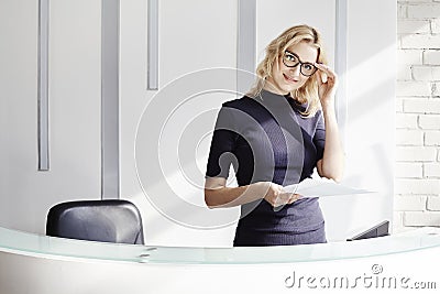 Beautiful blonde friendly woman behind the reception desk, administrator talking by phone. Sunshine in modern office. Stock Photo