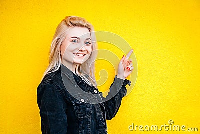 Beautiful blonde girl in black jacket smiling while lifting a finger up posing against a background of yellow wall. Copy, empty Stock Photo