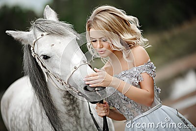 Beautiful blonde girl in dress strokes a gray horse on nature in Stock Photo